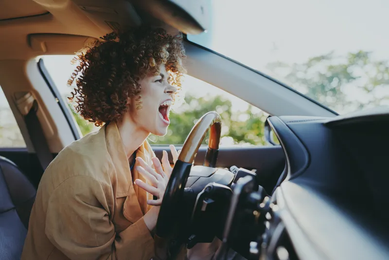 Curly haired woman screams in the driver seat, gripping the steering wheel as surprise shines through, a candid moment with a retro film color mood.