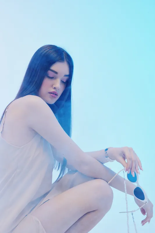 Young woman with long dark hair in a light dress poses holding sunglasses against a bright blue background. Concept of beauty, fashion, and summer style with soft natural lighting.