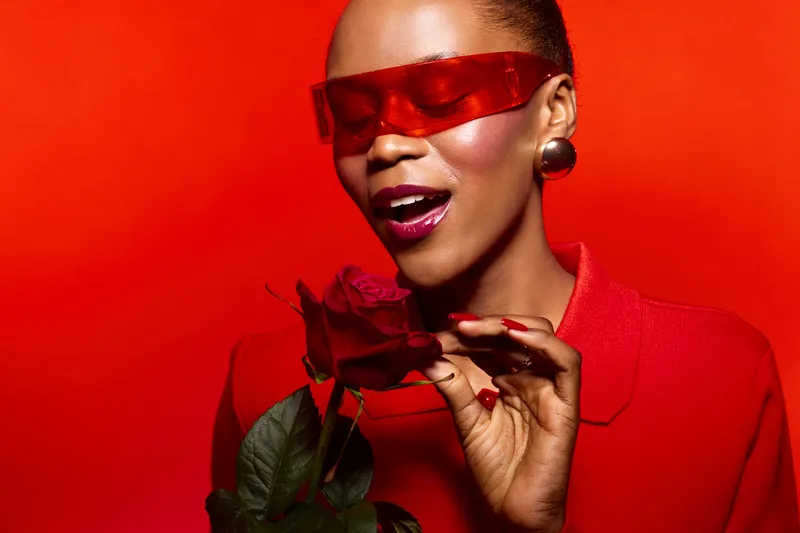 Stylish African American woman with dark skin wearing red sunglasses and red outfit holds red rose, posing against bright red background. Concept of fashion, beauty, and elegance.