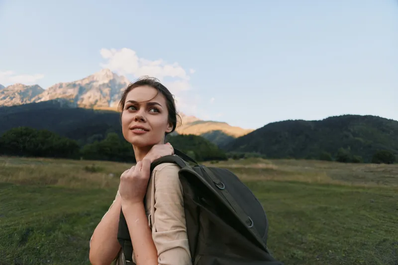 Young woman with a backpack walks through a green meadow toward distant mountains, feeling calm and curious during an outdoor adventure under clear skies