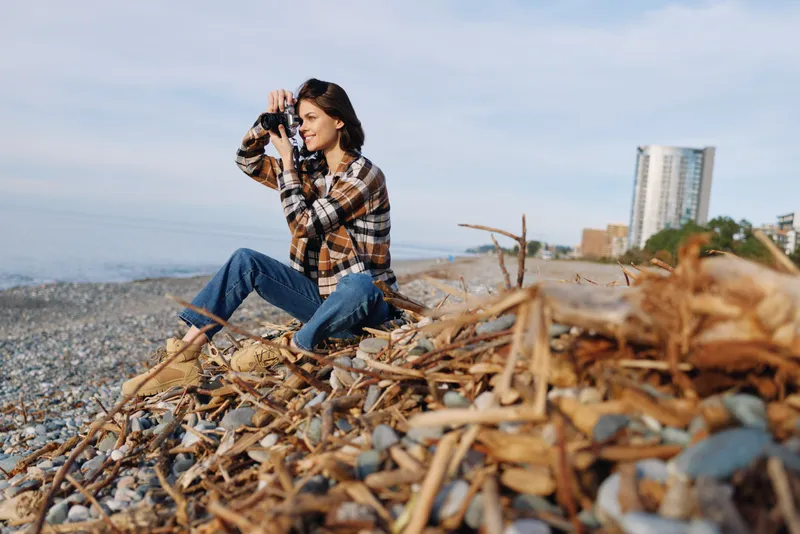 Woman on a beach with a camera, photographing the coastline amid seaweed, casual outdoor style, exploring nature and travel vibes.