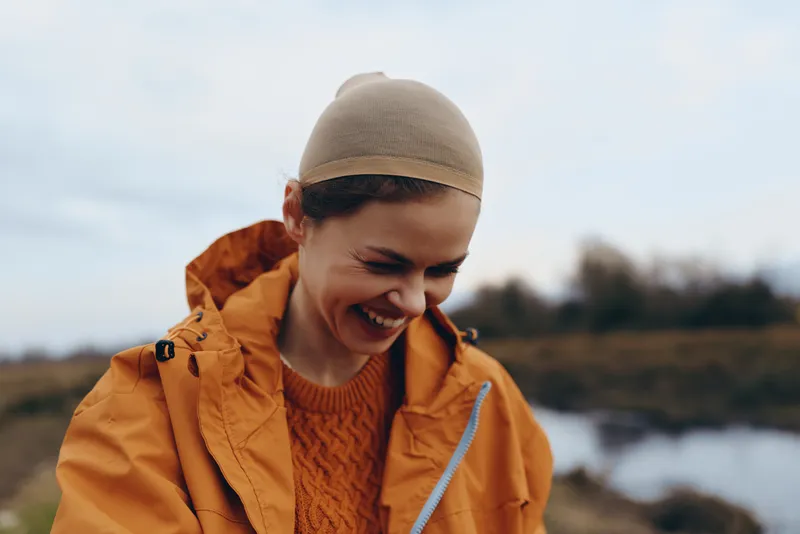 Woman lifestyle outdoors smiling in orange jacket and knit hat near lake path in soft daylight gentle autumn mood relaxed candid moment closeup profile windy air golden light today