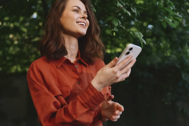 Young woman outdoors, smiling while holding a smartphone, casual red shirt, sunny greenery background, candid lifestyle moment, relaxed posture, natural colors, fresh vibe