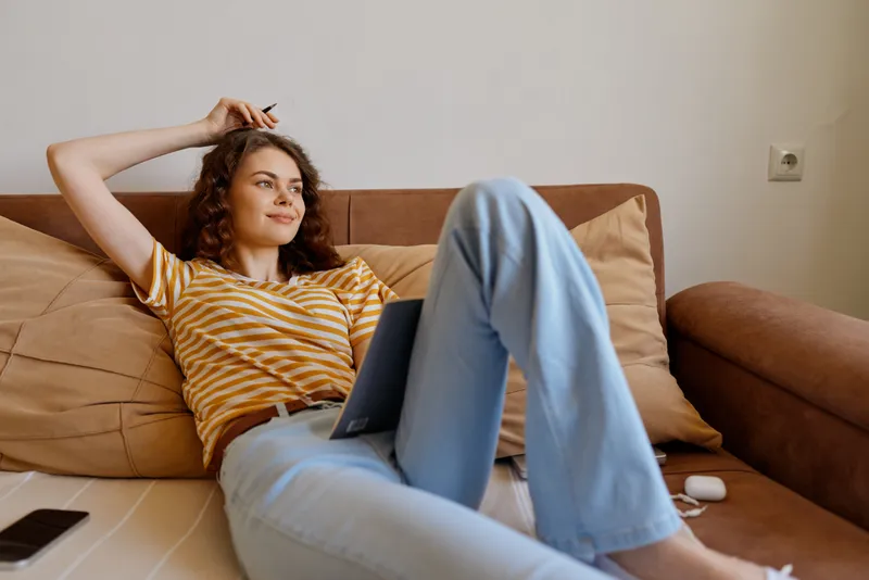 Cheerful woman at home, lounging on a sofa with a laptop, relaxed pose, warm lighting, casual striped shirt, soft furnishings, modern interior, content expression, lifestyle concept