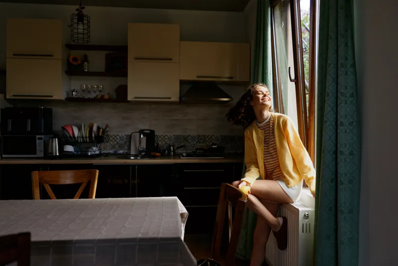 Woman relaxing in a sunny kitchen, sitting on a radiator ledge, wearing a yellow cardigan and striped dress, casual domestic scene capturing an intimate home atmosphere, natural light through window.