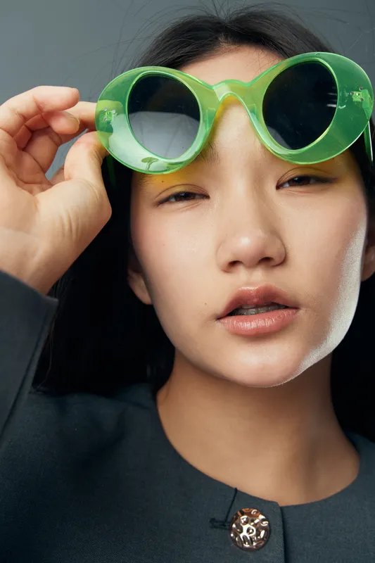 Woman closeup portrait with green sunglasses touching frame and looking at camera, natural makeup and black clothing, beauty and fashion concept with modern style.
