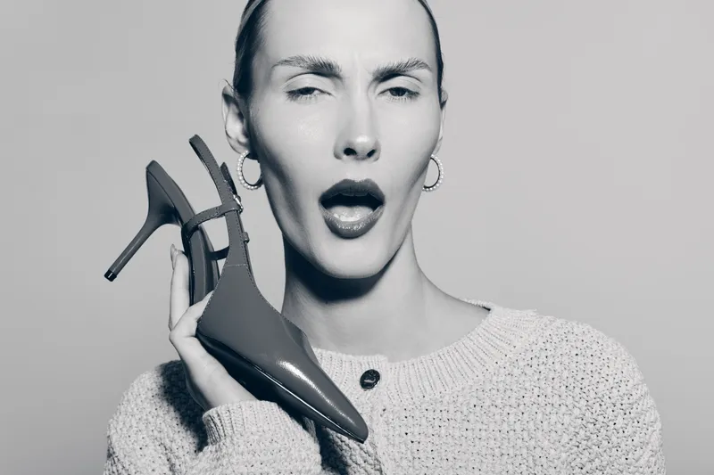 A slim Caucasian woman with light skin holds a high heel shoe near her face, expressing surprise with eyes closed and mouth open. Studio portrait on grey background.