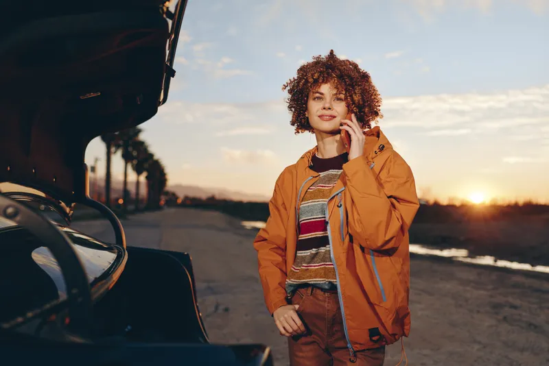Woman lifestyle moment by car on road at sunset, jacket and denim outfit, curly hair, standing outdoors with relaxed smile, open trunk beside vehicle, travel mood.