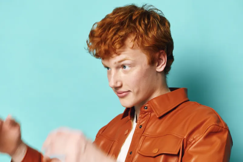 Young man with red hair wearing a brown leather jacket posing against blue background. Casual portrait of male model with focused expression and styled hairstyle in studio lighting.