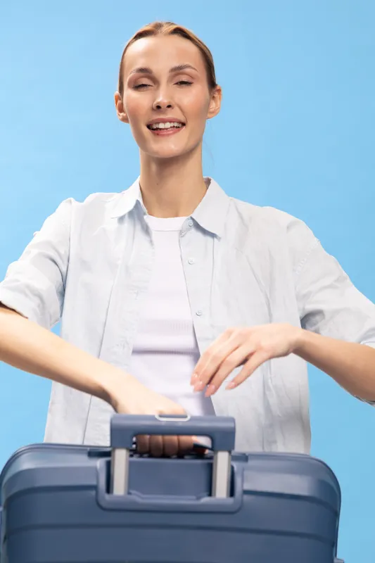 Young woman smiling while packing blue suitcase. Casual outfit and light background create cheerful mood. Concept of travel preparation and vacation planning.