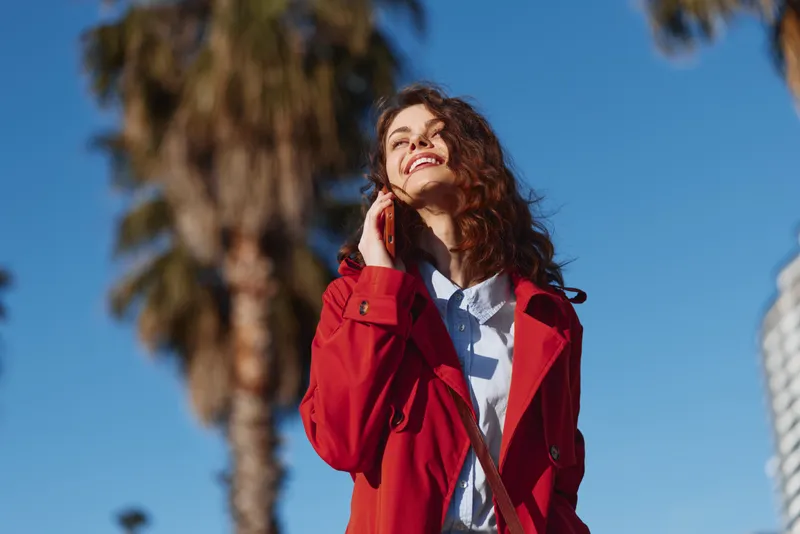 Young woman with curly hair smiles joyfully while talking on the phone outdoors wearing a red coat under clear blue sky near palm trees.