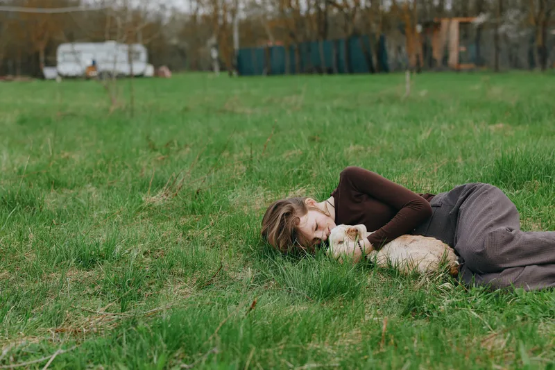 Relaxed woman lying on lush grass, gently cuddling a playful goat kid in a sunlit meadow, conveying calm, nature, and companionship, lifestyle moment in the countryside