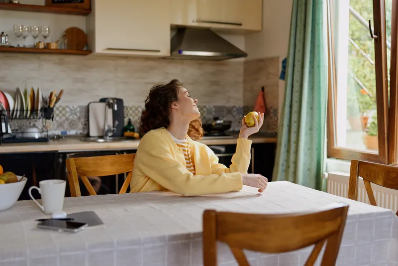 Woman at home in a bright kitchen, casual and relaxed, bathed in sunlight by a window, smiling over a morning orange, cup and tablet on the table, cozy atmosphere, warm colors, everyday lifestyle