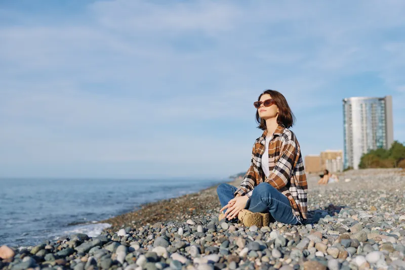 Woman sits on a pebbled beach by the sea in a plaid shirt and jeans, savoring calm coastal scenery, sunny weather, and a relaxed pose against a distant skyline at the edge of shore.