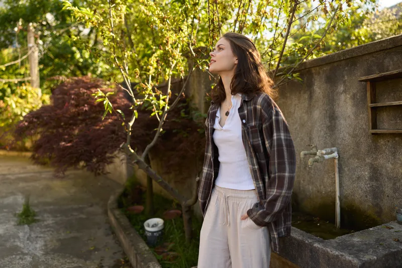 A young woman in a plaid jacket stands in a sunlit garden, hands in pockets, gazing upward with a contemplative mood. The casual outdoor setting and soft light convey a relaxed lifestyle moment