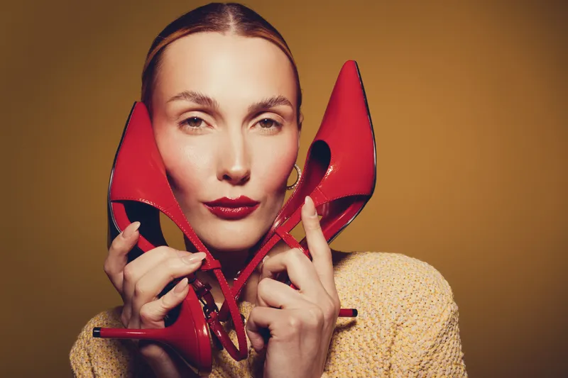 Stylish woman holds red high heel shoes near face, wearing yellow sweater. Fashion concept with bold makeup and confident expression on warm brown background.