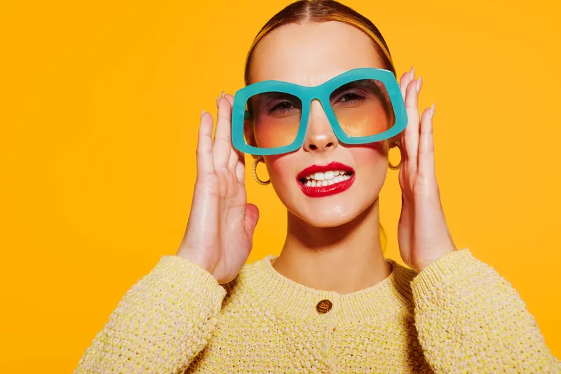 Stylish woman with bright red lipstick wearing large blue sunglasses and yellow sweater poses against vibrant orange background. Concept of fashion, summer style, and confident attitude.