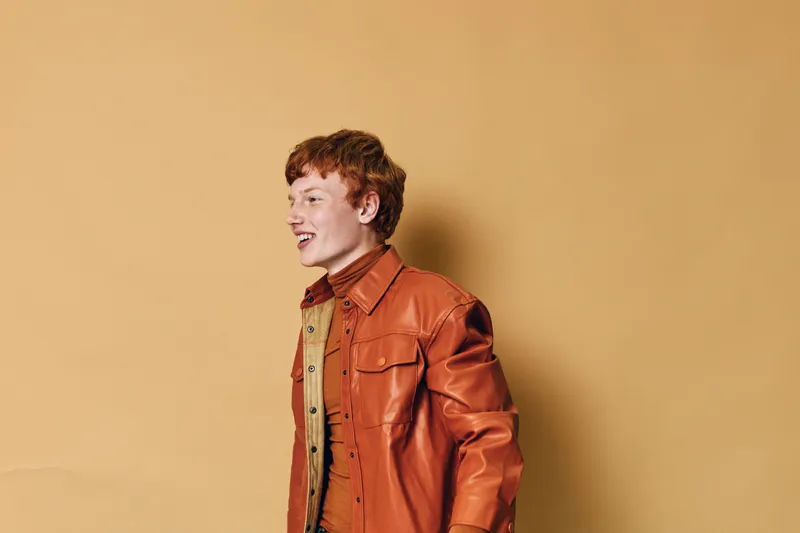 Redhead teenager smiling and looking aside with excitement wearing brown jacket and shirt against beige background. Youthful expression with casual fashion and side profile of boy.