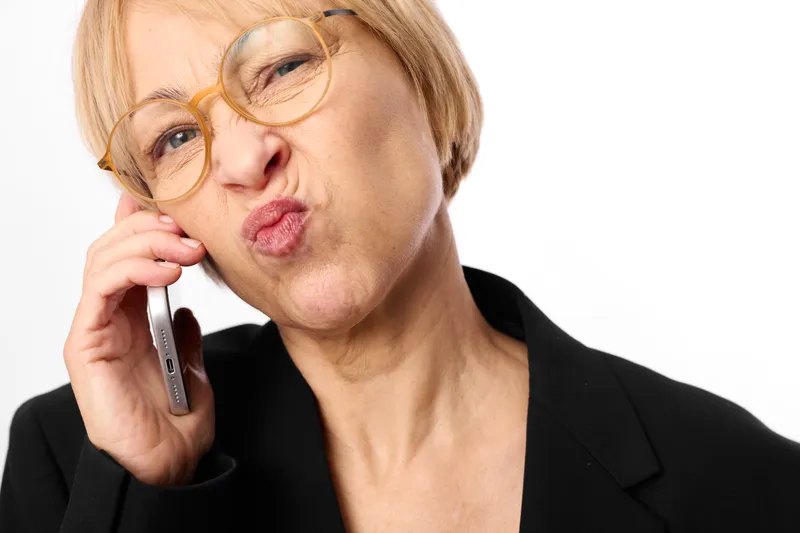 A worried senior woman with glasses holds a phone to her ear in business clothing, showing stress and anxiety on a clean white studio background, concept for communication and aging