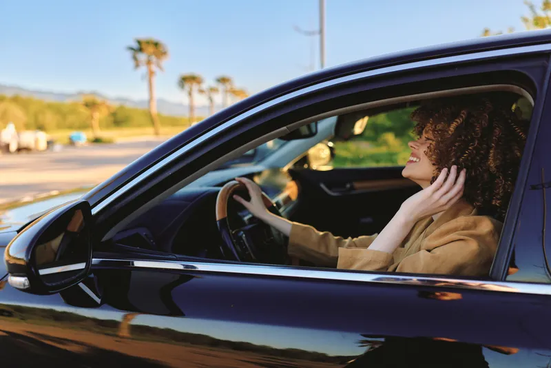 Morning light on a smiling woman driver behind the wheel of a sleek car, driving along a sunlit road. A casual road trip vibe with retro film color and warm tones. Concept: everyday lifestyle and