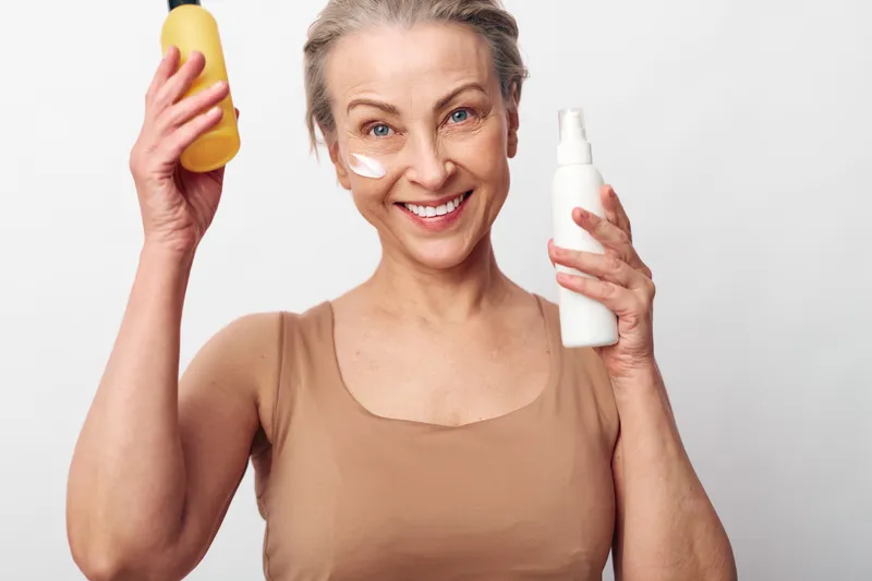 Mature woman with short grey hair smiles holding yellow and white skincare bottles, cream applied on cheeks. Bright studio background. Concept for skincare, beauty routine, aging skin care.