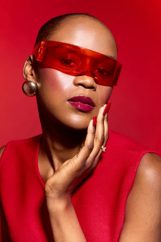 Portrait of black woman with dark skin wearing red translucent glasses and red outfit, posing with manicured nails and bold lipstick on red background. Fashion and beauty concept.