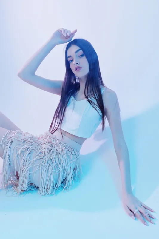 Young woman beauty fashion model poses sitting on the floor in stylish outfit with long hair in soft blue studio lighting. Concept for modern style and elegance.