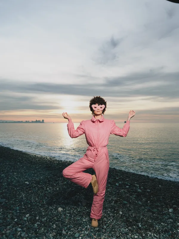 Beach scene at sunset with a woman in a pink jumpsuit performing a balanced yoga pose on pebbles along the coast, calm ocean in the background and a serene lifestyle moment
