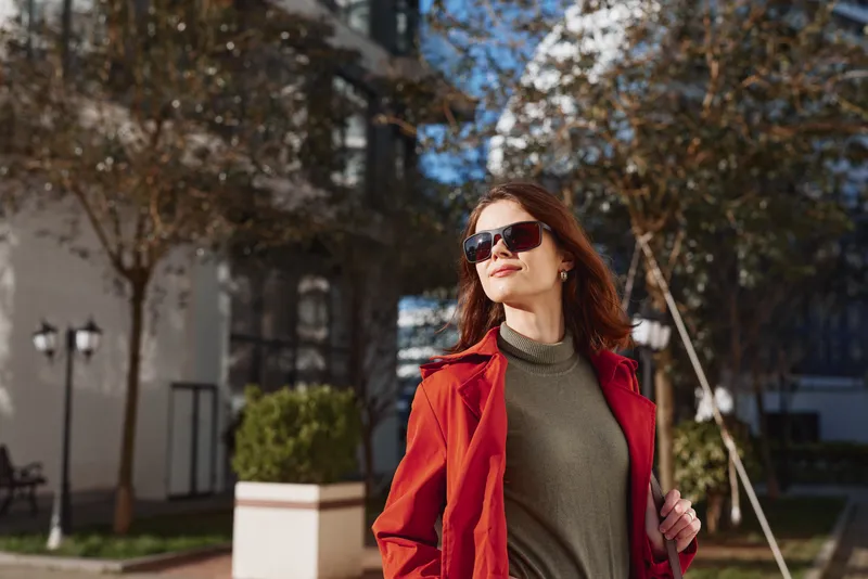 Confident young woman in a red coat and olive sweater walks outdoors under bright spring light, posing with effortless style and focus, her expression calm and assured.