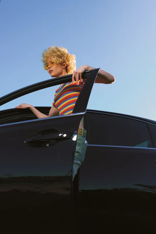 Cheerful woman outdoors, leaning from a sleek car with a striped top, sunlit moment, relaxed pose, soft breeze, vintage tones, cinematic lifestyle snapshot, blue sky and road vibrant.