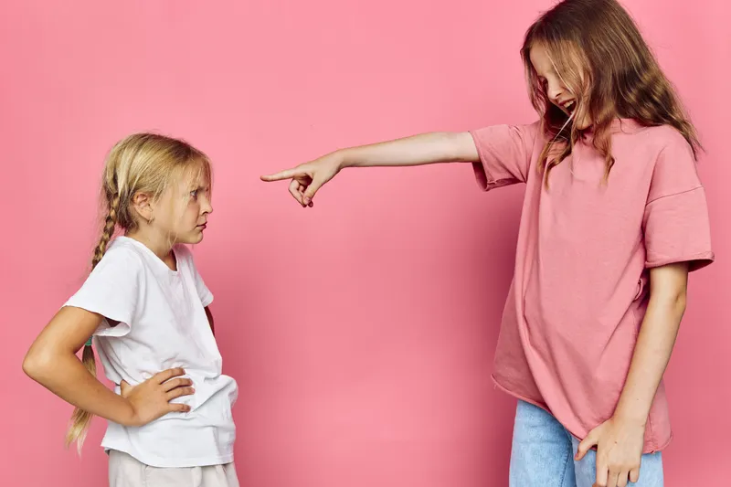 Two young girls stand against a pink background while one points and shouts at the other with a stern expression.