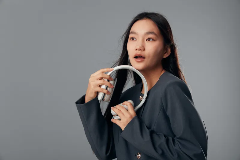 Young woman with long black hair wearing dark blazer holds headphones near face in thoughtful expression on gray background. Studio portrait of female with gadget in hand for leisure.