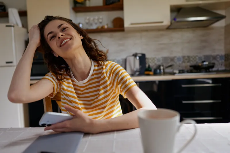 Young woman with curly hair sits at a bright kitchen table, smiling as she taps on a smartphone.