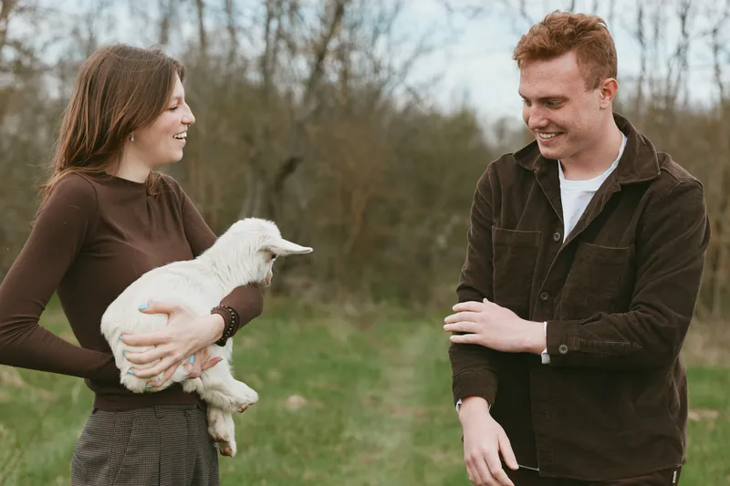 Cheerful woman outdoors, holding a white goat kid, smiling with a nearby man, warm natural light, relaxed mood, casual clothing, gentle breeze, countryside backdrop, wholesome moment