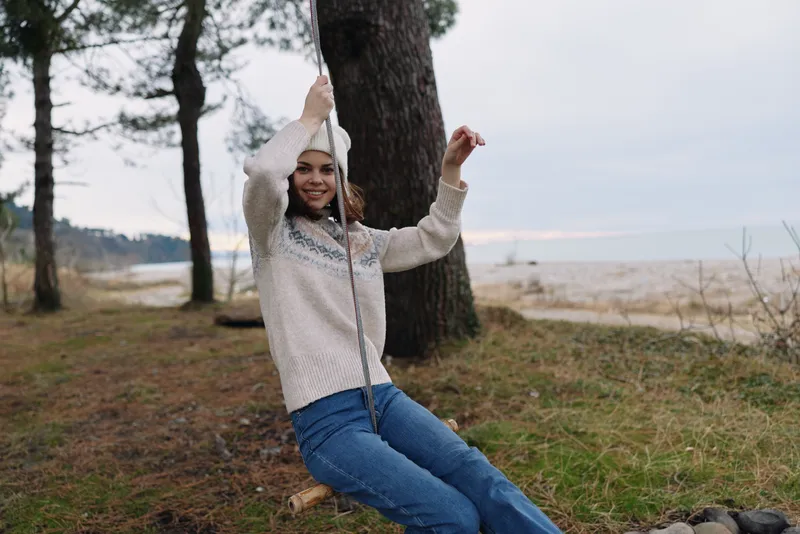 Girl on a swing in a peaceful outdoor setting by the shore, wearing a cozy sweater and jeans, with trees framing the scene and a calm sky for a serene nature moment.