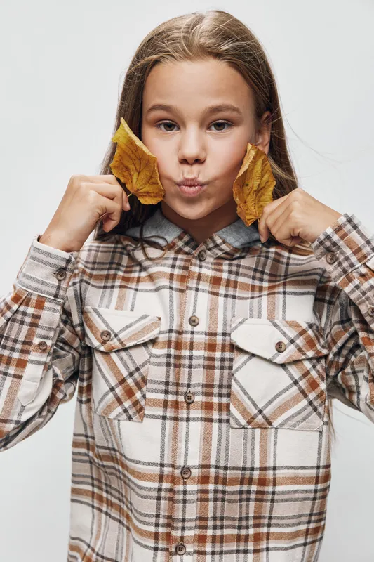 Girl with long hair holding yellow autumn leaves near face wearing brown checkered shirt on light background in studio for fall season and childhood concept.