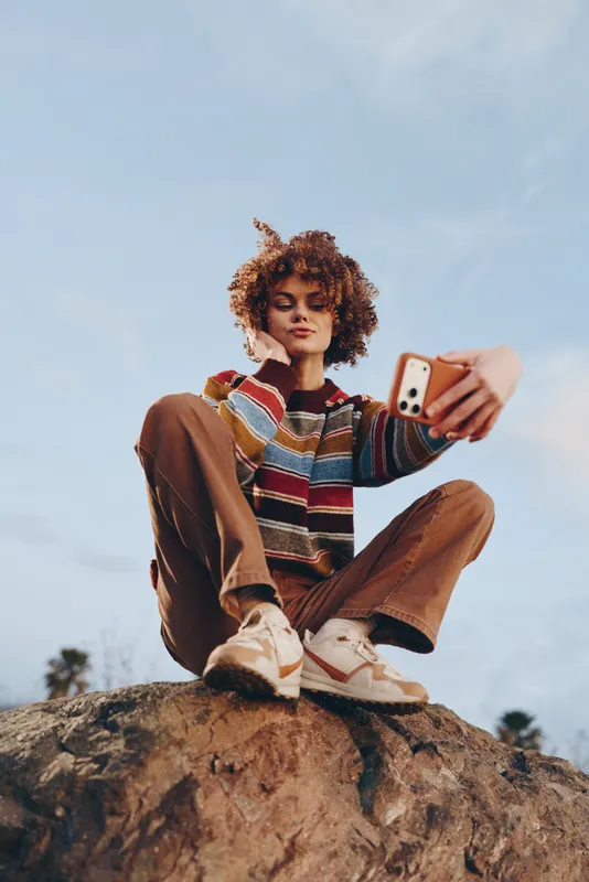 Woman lifestyle outdoors, smiling softly while taking a selfie on a sunlit rock, curly hair blowing in the breeze, casual sweater and jeans, holding a smartphone, blue sky, open air