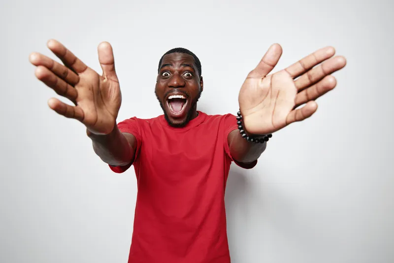 African man with dark skin wearing red t shirt smiling excitedly with open arms and wide eyes on white background for happiness and welcome gesture.