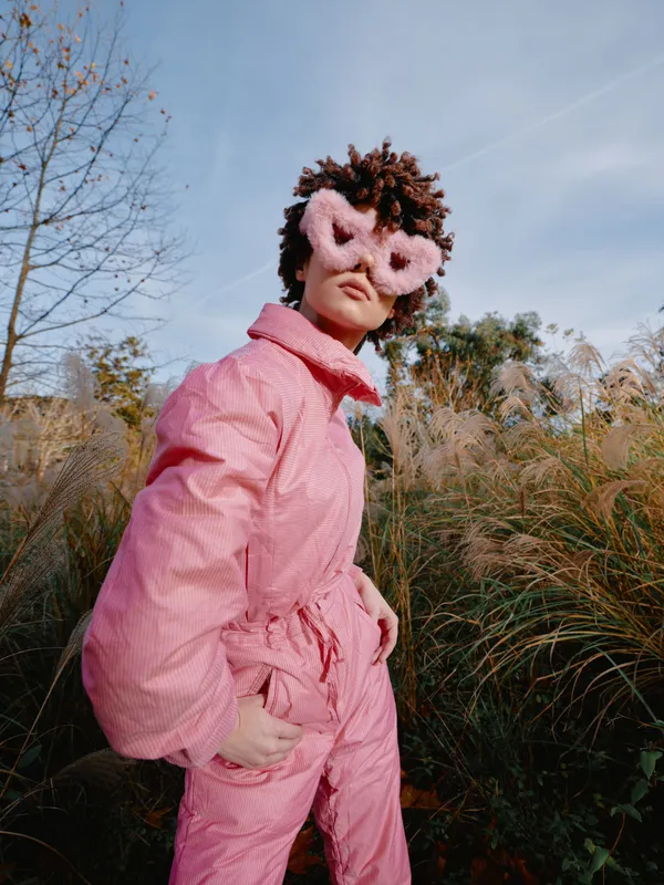 Person in a pink jumpsuit and fluffy heart-shaped mask poses outdoors among tall grasses under a clear sky, creating a whimsical fashion moment with nature as a backdrop.