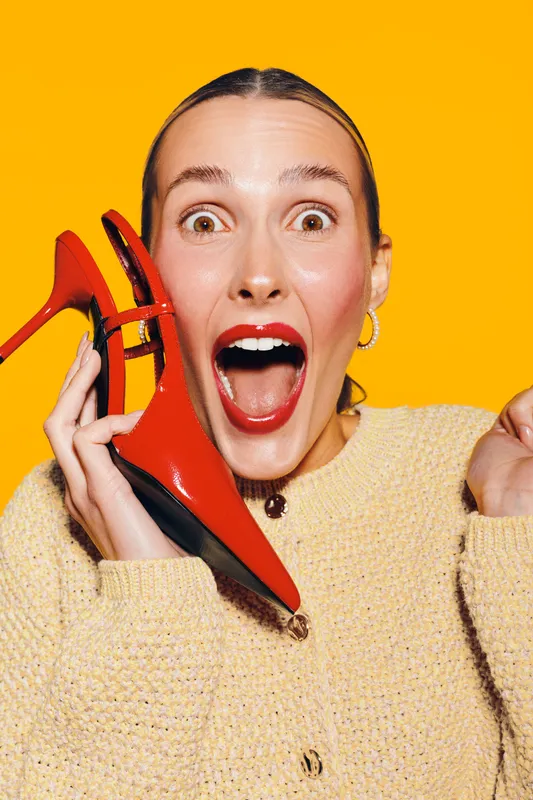 Excited woman with red lipstick holds red high heel shoe near face, wearing beige sweater on bright yellow background. Concept of fashion, style, and joyful expression.