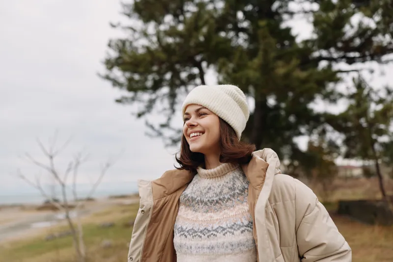 Woman outdoors in winter gear smiling, wearing a beanie and puffer jacket over a knit sweater, beside trees by the water in a scenic park.
