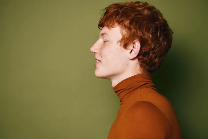 Portrait of a young man with red hair and freckles wearing a brown turtleneck sweater against green background. Side view with closed eyes and relaxed facial expression.