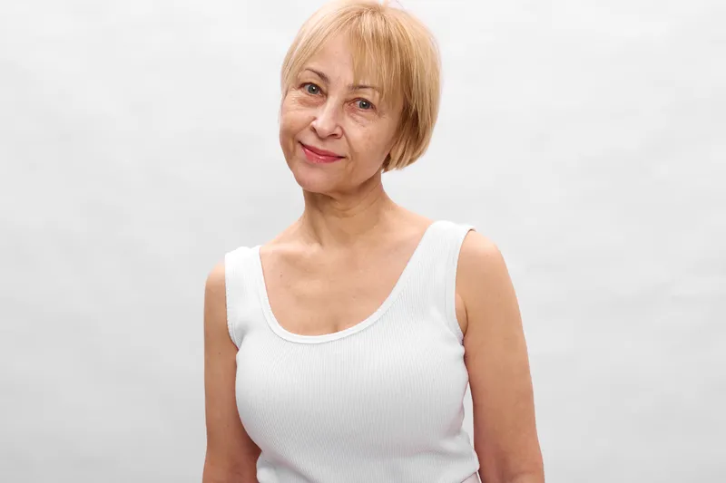 Senior woman elderly mature, smile, white studio portrait, aging wellbeing, short blond hair, sleeveless white top, calm expression, white background