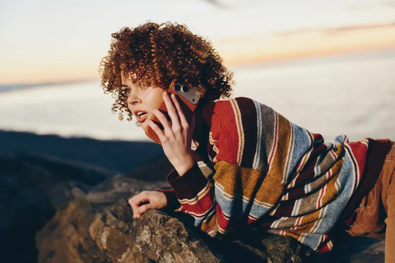 woman lifestyle portrait outdoors phone communication seaside curly hair sweater, woman talks on mobile near rocky coast, relaxed smile, warm striped poncho, natural sunlight