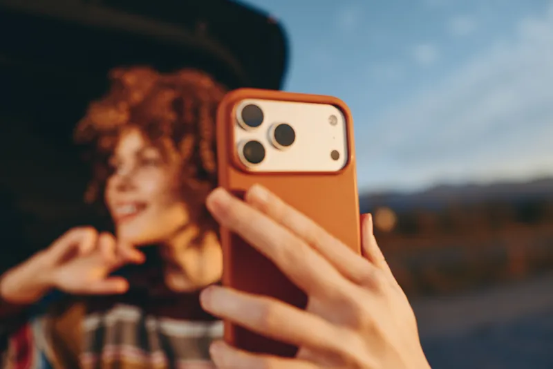 Woman lifestyle selfie outdoors, closeup hand holding smartphone with orange case, curly hair and relaxed smile, mountains scenery, blurry background, evening light, travel mood