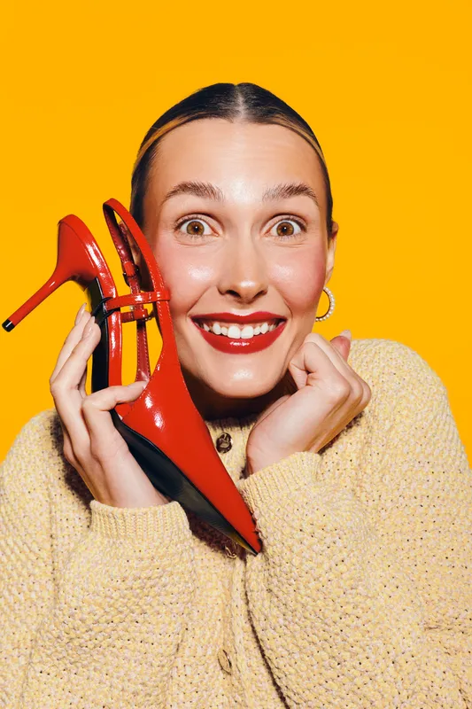 Happy woman with red lipstick holds red high heel shoes near face on yellow background. Concept of fashion, style, beauty, and joyful emotions.