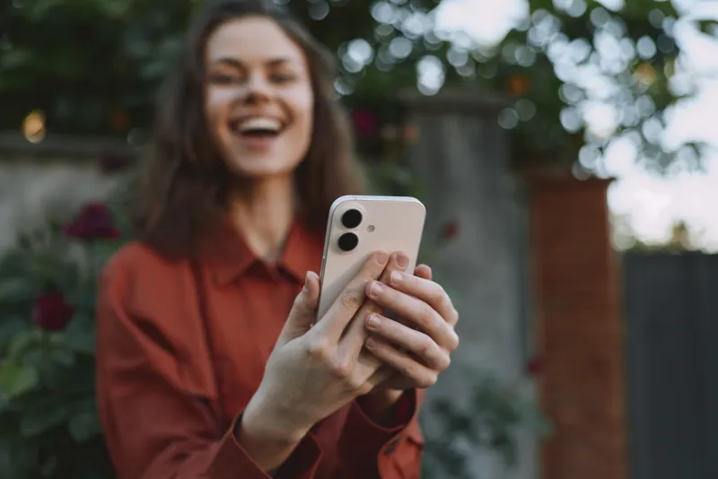 Woman smiles while taking a selfie outdoors, holding a smartphone in front of a lush garden. Casual lifestyle moment captured in natural daylight with a candid, relaxed pose