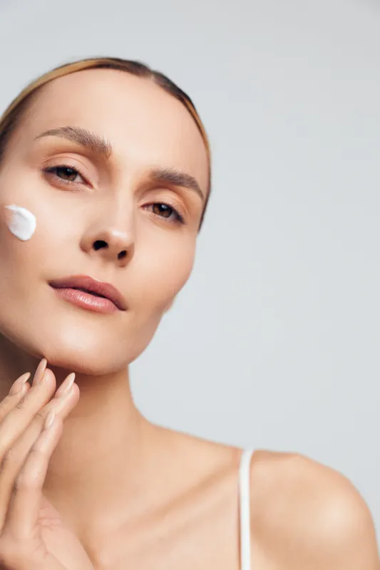 Woman with clear skin applying white face cream on cheek, close-up portrait on light background. Concept of skincare, beauty routine, self-care, natural complexion, wellness.