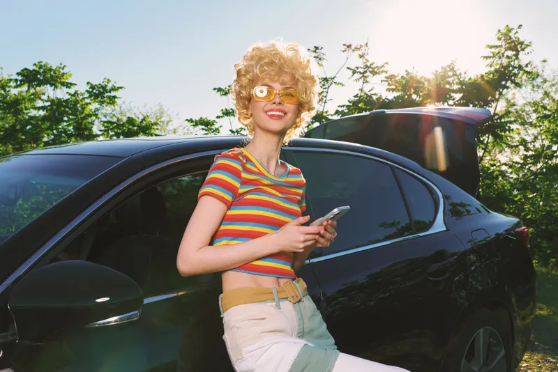 Cheerful woman outdoors, striped shirt, posing beside a dark car, sunlit moment, relaxed stance, soft breeze, vibrant colors, natural greenery background, candid lifestyle mood, warm summer atmosphere
