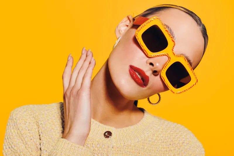 Stylish woman with slicked hair wearing oversized orange sunglasses and red lipstick poses against bright yellow background. Fashion concept with bold colors and modern accessories.